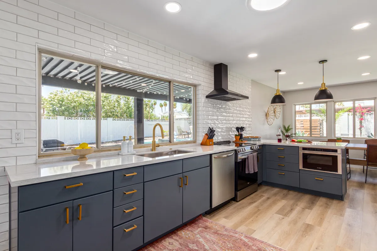 A contemporary kitchen featuring a spacious kitchen island, large window bathing the room in natural light, sleek stainless steel appliances, and decorative wall accents. The scene is styled for gathering and entertaining, with ample workspace and inviting ambiance.