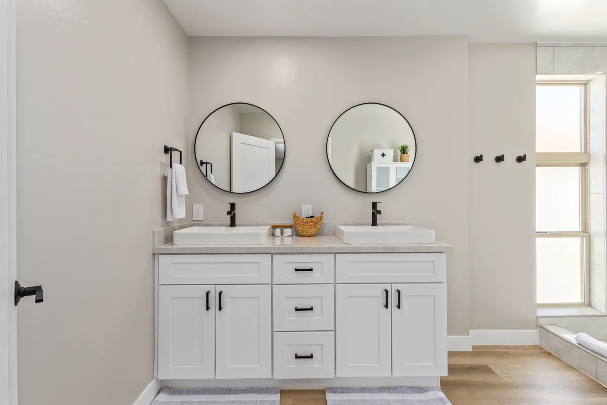 A spacious bathroom featuring double sinks with circular mirrors, bathed in natural light from a large window. The neutral palette and clean lines evoke a spa-like ambiance, perfect for refreshing morning routines or relaxing evening rituals.