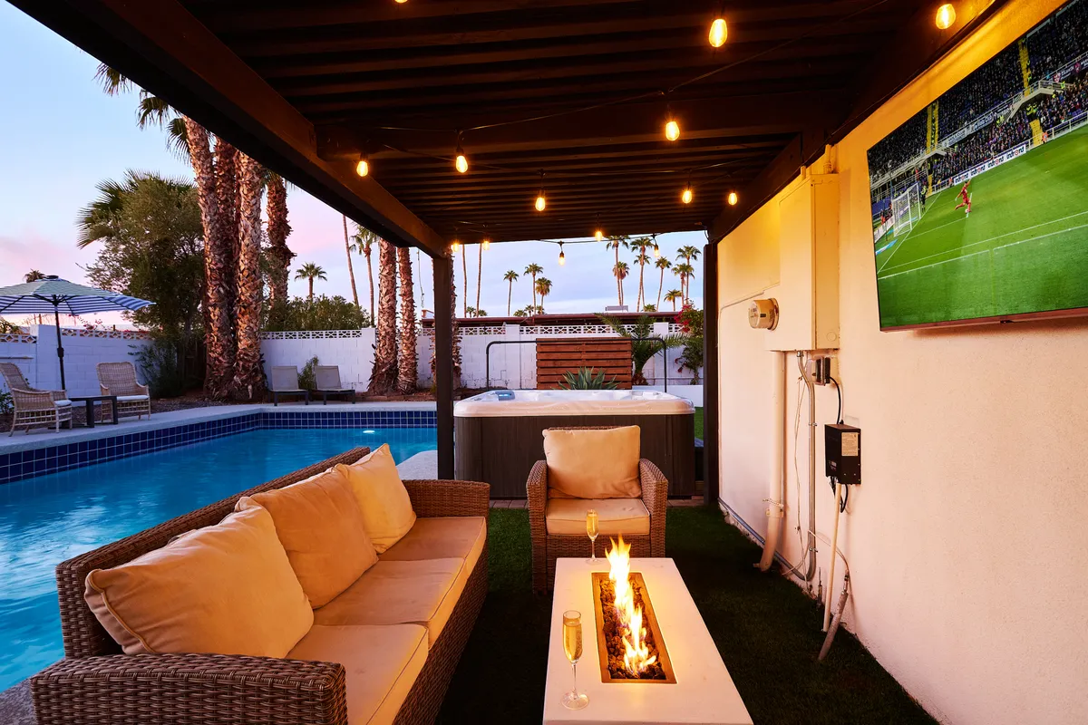 A stylish backyard setting featuring a fire pit surrounded by comfortable seating, a hot tub and pool in the background, and a large TV screen perfect for outdoor sports or movie nights. The area is designed for relaxation, group gatherings, and festive socializing.