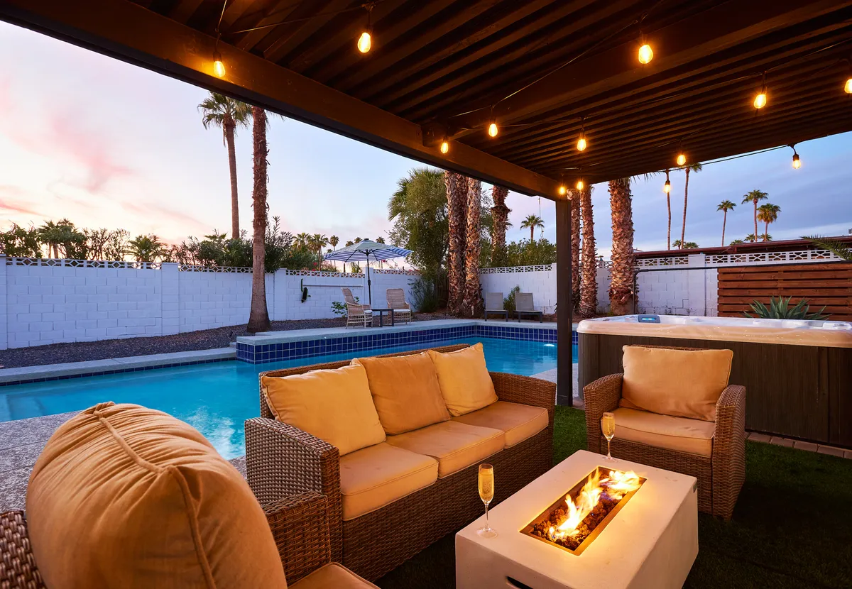 A cozy backyard scene with a glowing fire pit surrounded by comfortable seating, the pool and hot tub nearby, and lush palm trees setting a tranquil tone. The area feels inviting for both daytime and evening relaxation or socializing.