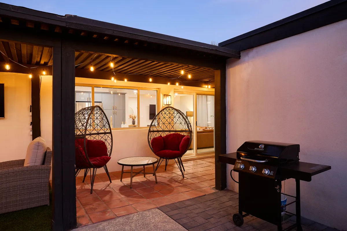 A cozy patio setting featuring hanging egg chairs, a round coffee table, and a barbecue grill, with string lights creating a warm ambiance. The layout encourages outdoor dining, conversations, and relaxed social gatherings as the sun sets.