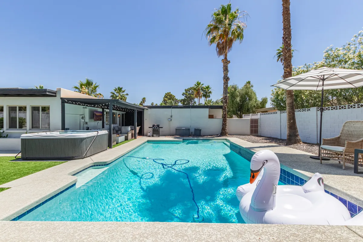 A lively backyard view featuring a sparkling swimming pool with a floating swan, an adjacent hot tub, palm trees, and a stylish outdoor seating area. The setup hints at sunbathing, swimming, and cheerful gatherings, with the sunset casting a warm glow over the scene.