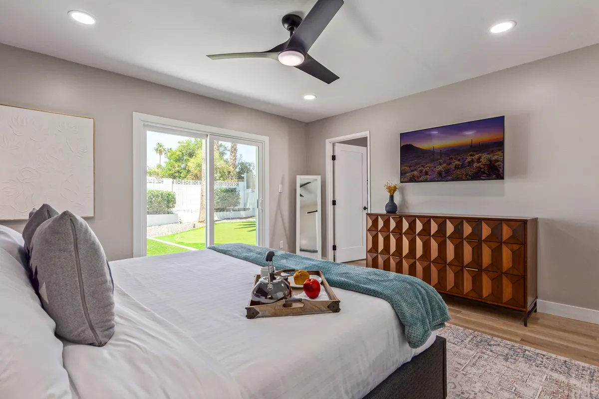 A serene bedroom featuring plush bedding and pillows on a comfortable bed, a stylish wooden dresser with geometric design, large windows letting in abundant natural light and offering a view of the garden, and a modern ceiling fan overhead. The tranquil setting is ideal for relaxation, with space to enjoy coffee in bed or prepare for the day in comfort.