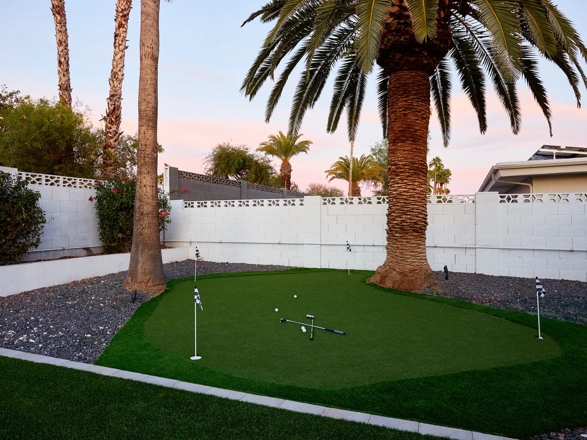 A private backyard putting green framed by palm trees, a white privacy wall, and thoughtfully landscaped stones. The tranquil space is bathed in soft evening light, perfect for practicing short game skills or relaxing outdoors at home.